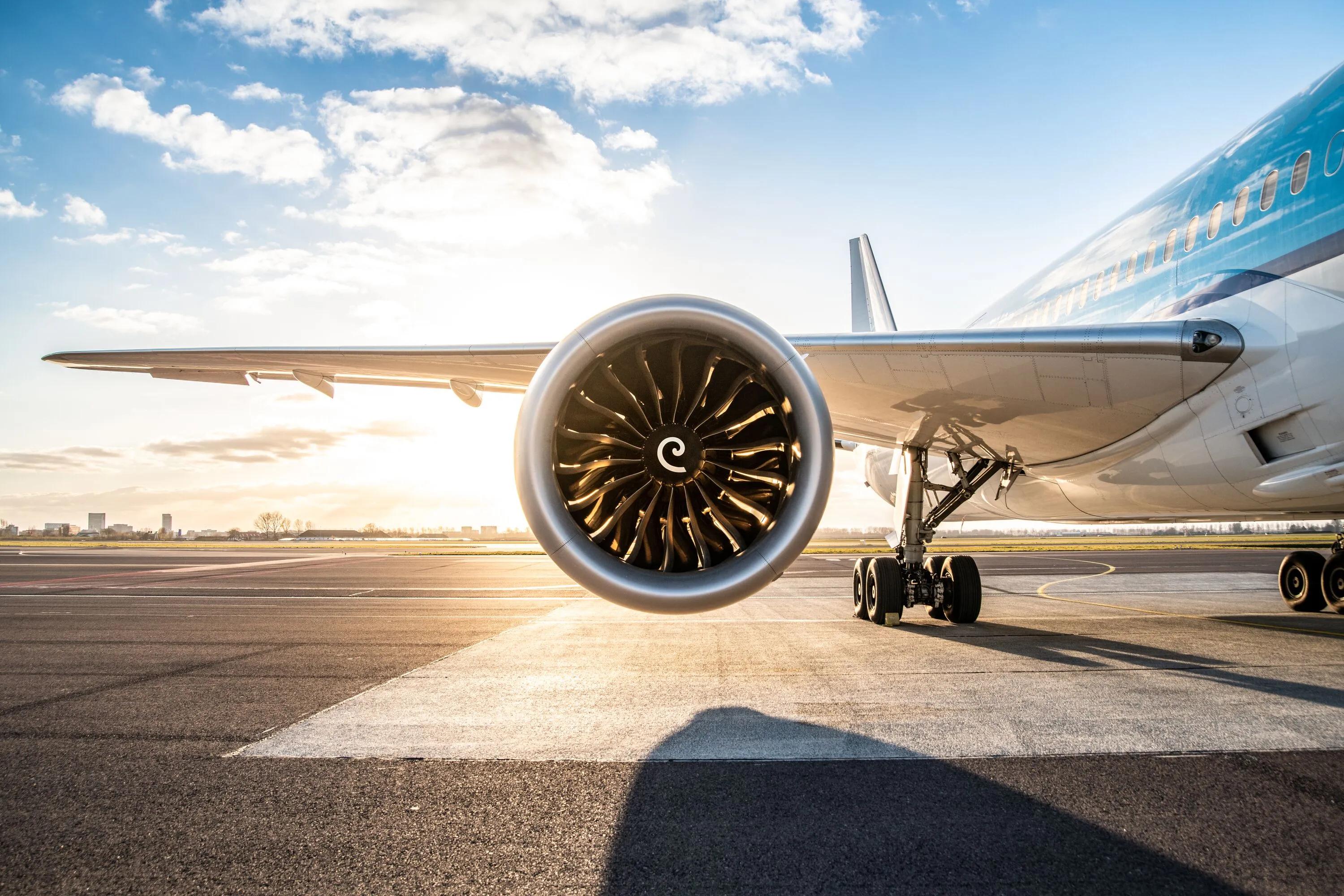 Low-angle view of a KLM airplane on the runway, with a large jet engine in the foreground, landing gear beneath the wing, and warm sunrise light reflecting on the aircraft.